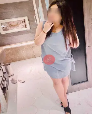 A woman with long dark hair stands in a modern bathroom, wearing a sleeveless gray dress and black high-heeled shoes. Holding her hair with one hand and slightly posing, she exudes an air of elegance akin to a call girl in Delhi. The bathroom boasts marble floors and a framed photograph on the wall.