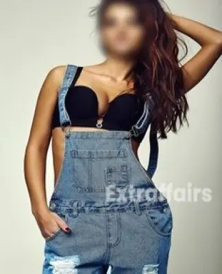 In a trendy studio, a call girl service provider with long hair poses wearing denim overalls and a black bra.