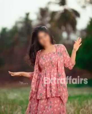 A woman wearing a pink floral dress is smiling and posing outdoors with her arms raised, capturing the beauty found near the lush greenery and trees, reminiscent of tranquil moments one might experience while exploring delightful spots Call Girl in Noida.