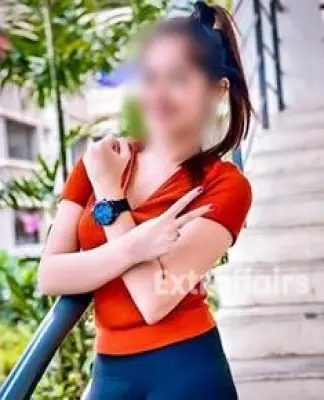 A young woman with a cheerful smile stands by a stair railing outdoors, perhaps just returning from a relaxing massage service in Noida. She wears a red top, dark pants, and a watch, with her hair tied back. Her right hand forms a peace sign, and lush greenery is in the background