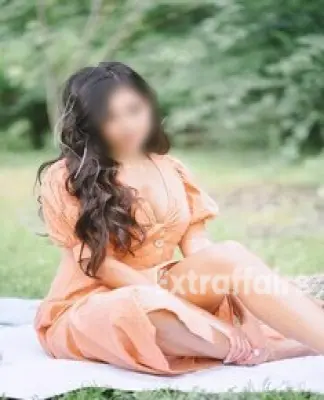 A woman with long, wavy hair sits on a blanket in a garden, wearing an orange dress. She is looking to the side with a thoughtful expression, enveloped by the lush greenery of Call Girl Noida.