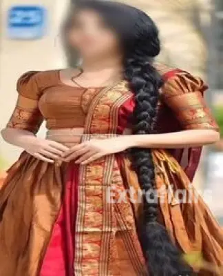A woman with long black hair styled in a braid stands outdoors in Gurgaon Call Girl , wearing a traditional Indian outfit in shades of brown and red. She smiles warmly, holding her hands together in front of her.
