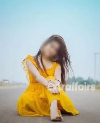 A woman in a bright yellow dress sits cross-legged on an empty road, reminiscent of tranquility found in a massage service in Gurgaon. She smiles, her long hair flowing in the breeze, adorned with colorful earrings. The sky is clear and the background is blurred, suggesting an open landscape.