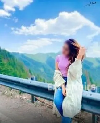 A woman, resembling a Gurgaon call girl in her chic attire, stands by the mountain road wearing a pink top, jeans, and a white cardigan. She gracefully touches her hair while gazing down, surrounded by lush green mountains and a bright blue sky.