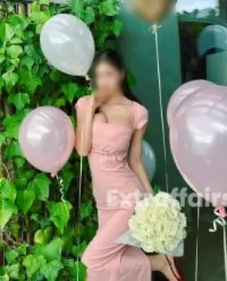 A Call Girl In Raj Nagar Extension in a pink dress poses with a bouquet of white roses, surrounded by pink and white balloons, reminiscent of the serene ambiance one finds during a full body massage in Raj Nagar Extension. She stands gracefully against a lush, leafy green background.